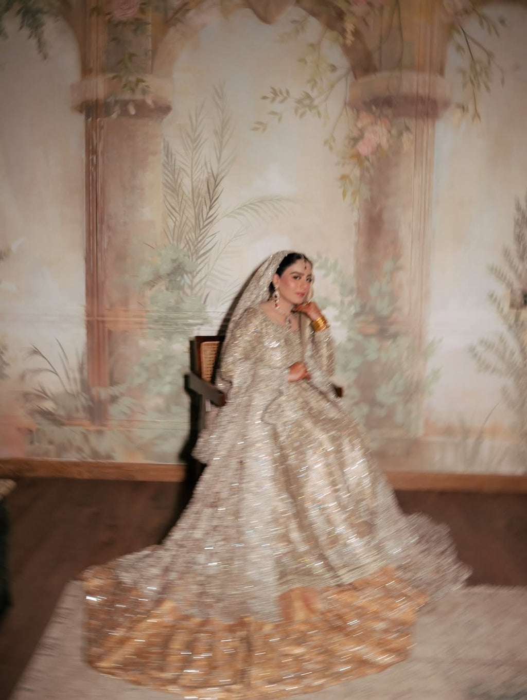 Bride wearing champagne silver embellished bridal gown with sweeping train and matching dupatta, seated in vintage mural backdrop - Èlaan Studio Pakistani wedding wear