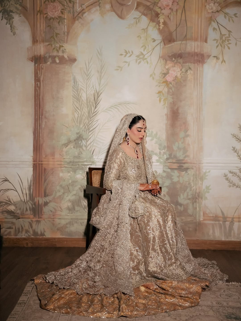 Bride wearing champagne silver embellished bridal gown with sweeping train and matching dupatta, seated in vintage mural backdrop - Èlaan Studio Pakistani wedding wear
