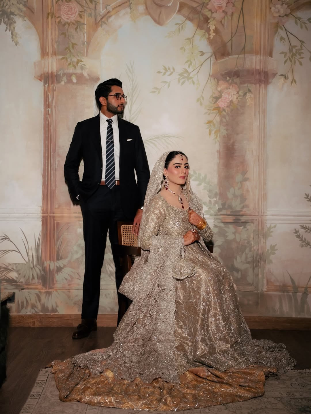 Bride wearing champagne silver embellished bridal gown with sweeping train and matching dupatta, seated in vintage mural backdrop - Èlaan Studio Pakistani wedding wear