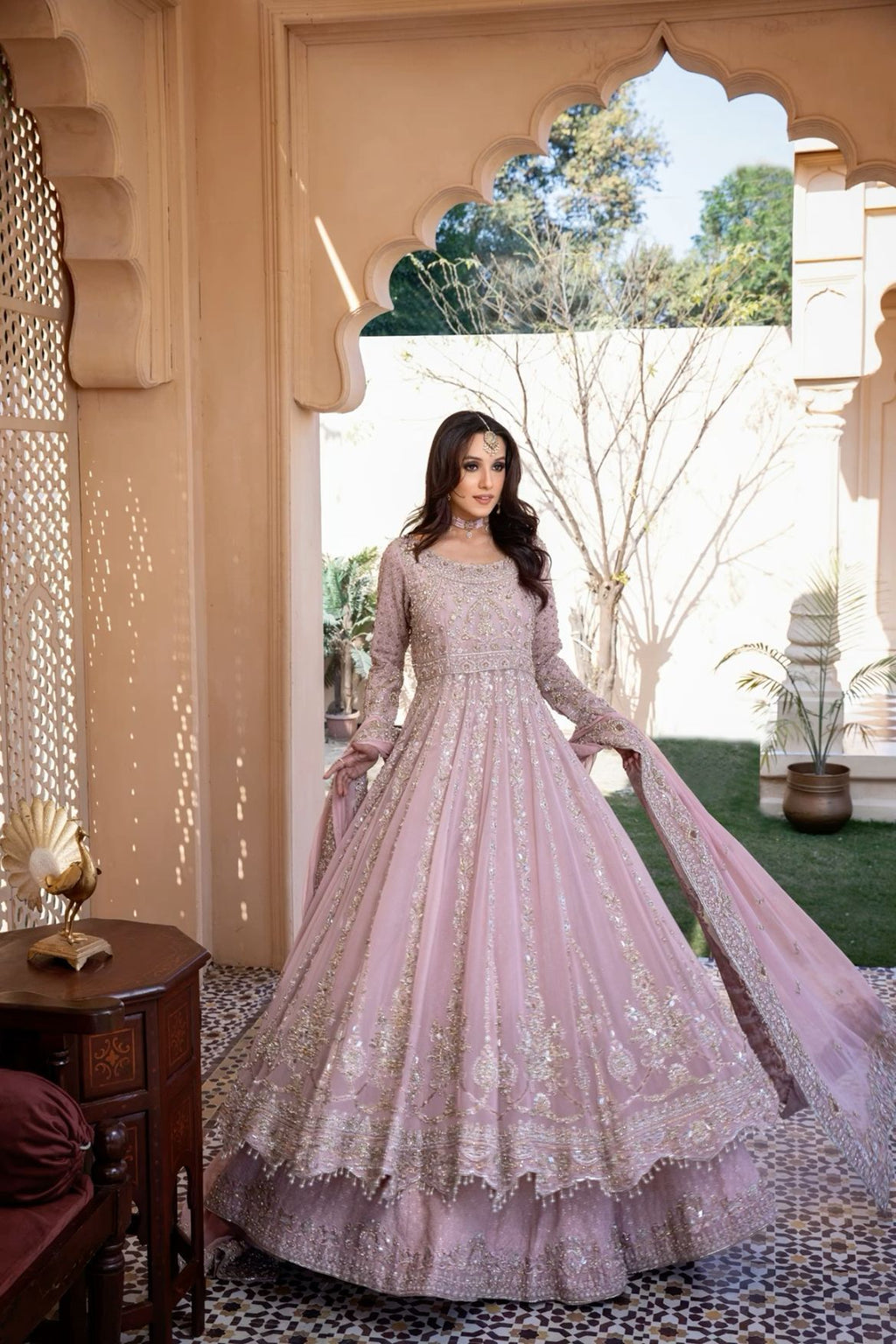 Model wearing blush pink embellished pishwas gown with silver dabka embroidery and matching dupatta, standing in Mughal-style archway - Èlaan Studio bridal couture
