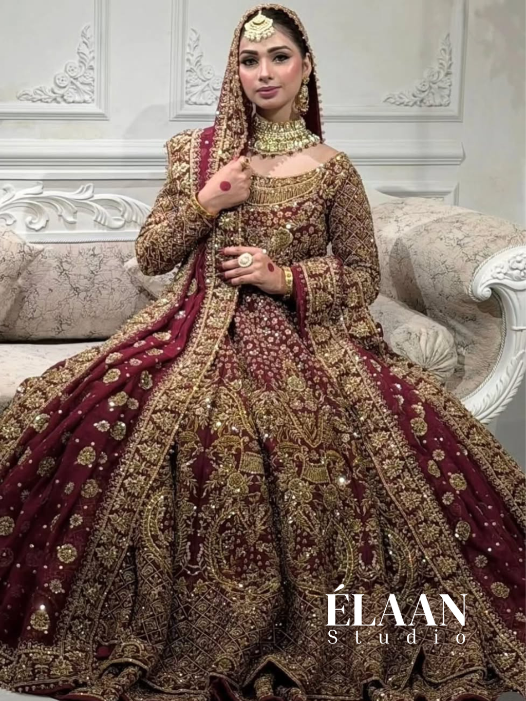 Bride wearing maroon gold heavily embellished bridal gown with embroidered dupatta, seated on ornate sofa - Èlaan Studio bridal wear