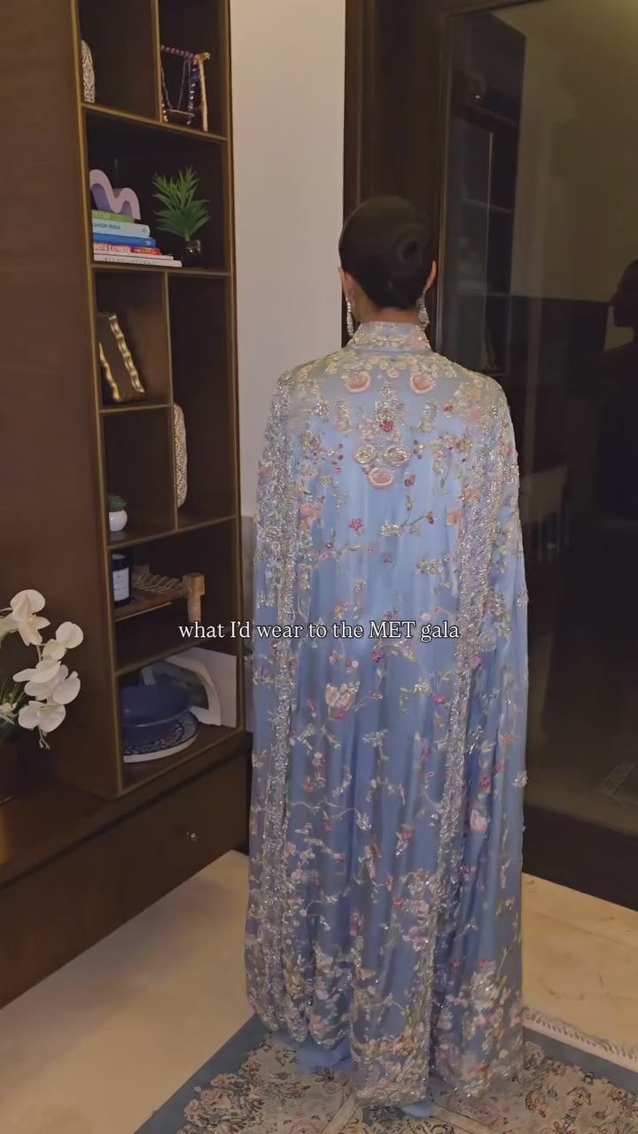 Woman in dusty blue floral embroidered cape, side view showing floor-length drape